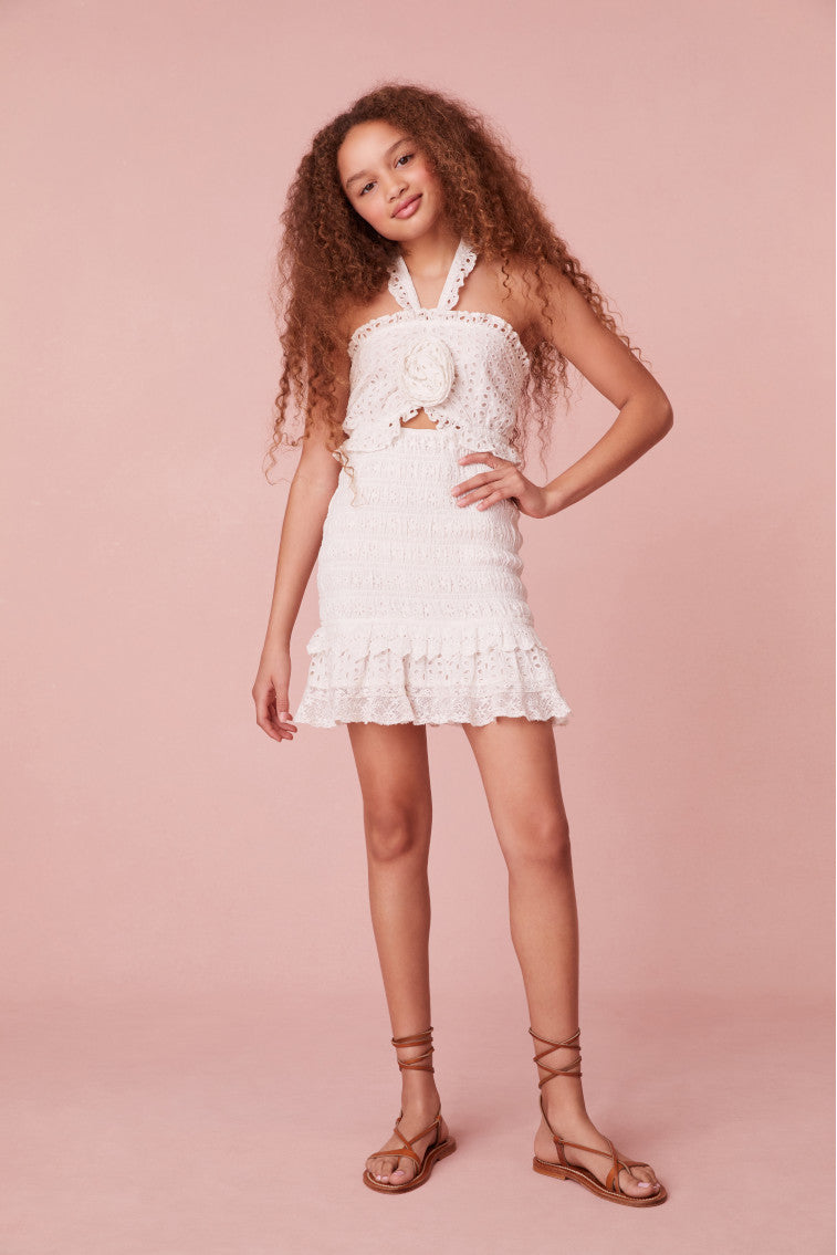 Girls mini dress with eyelet details all over the neckline, bodice, and hemline. A rosette sits at center front above a slight keyhole opening. The skirt is smocked throughout before falling to the two tiered ruffle skirt.