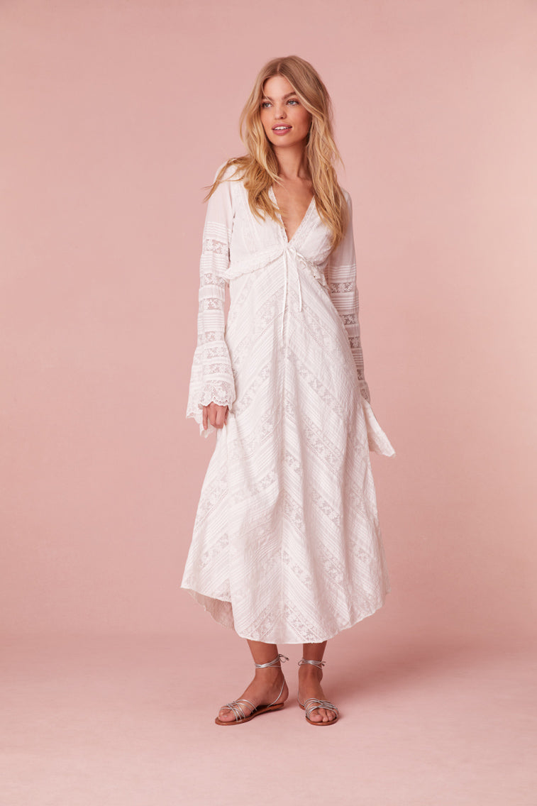 Maxi dress with long bell sleeves featuring a deep v-neck, a tiny ruffle at the waist that descends to a skirt with diagonal laces in a chevron design with textured pintuck panels. Features a zipper at the side seam.