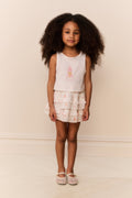 Young girl wearing a white sleeveless top and ruffled skirt with pink flower design on a beige background