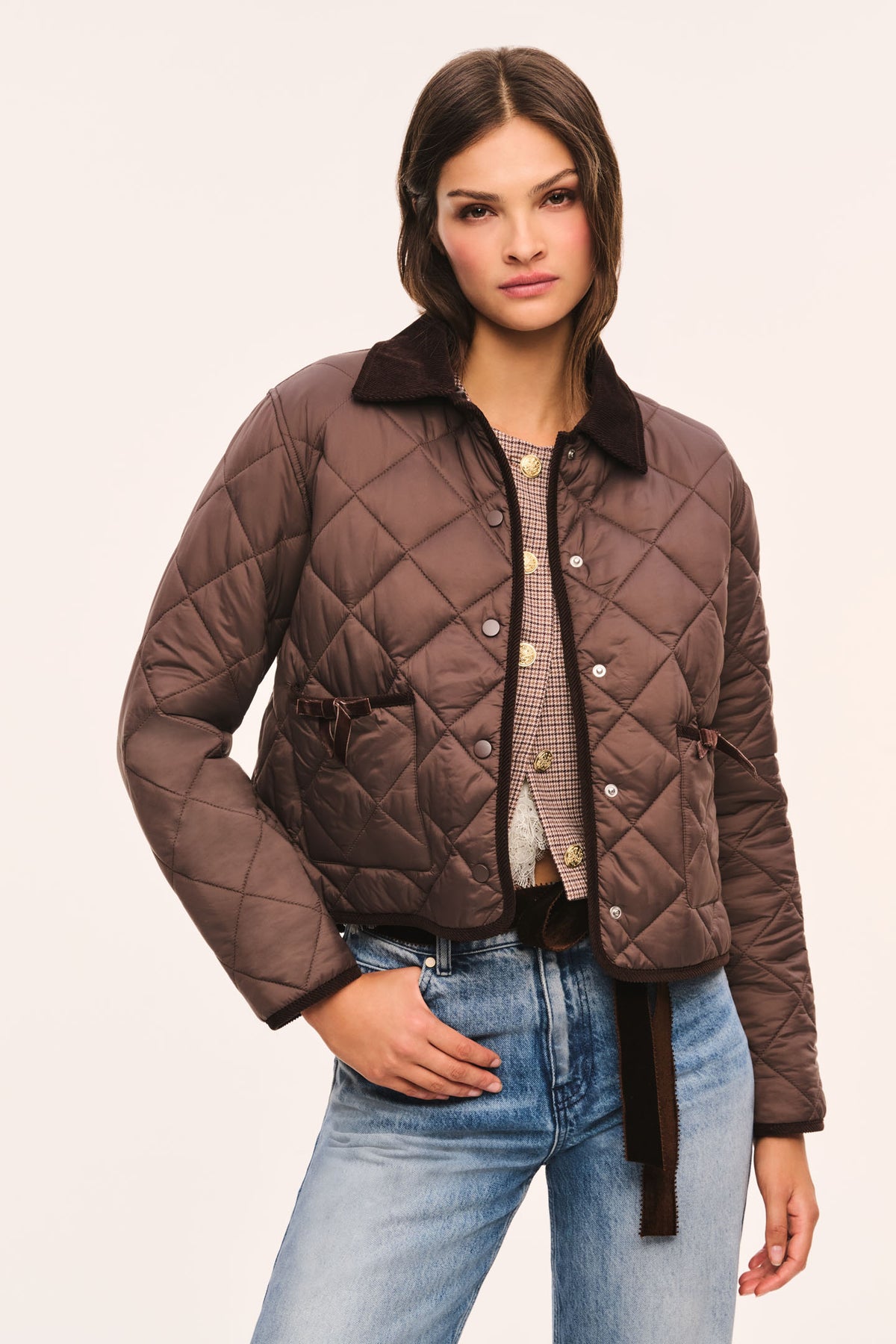Front view of brown quilted jacket with dark brown corduroy collar and corduroy trim, snap button front, and front pockets with velvet bow trim, and model with her hand in her pocket