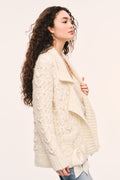 Windel ivory knit cardigan side view: ivory textured knit cardigan, open front with shawl collar, ribbon laced through the front, and ribbon bows on the pockets