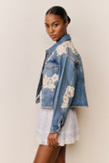 Woman wears blue denim jacket with floral lace embroidery and a light blue floral skirt on a beige background