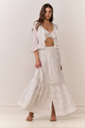 Woman wearing a white button down blouse with ruffled details and bralette top with matching maxi skirt on a plain background