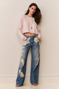 Woman wearing light pink sweater and blue jeans with floral lace appliqué against a beige background