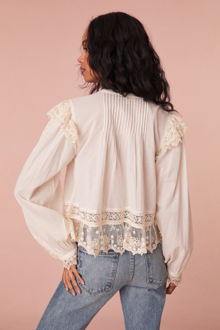 White long sleeve blouse featuring mesh and laces throughout. Has delicate mesh ruffles at the shoulder of the long blouson sleeves, shirred mesh ruffles with scallop edges at the hem, and a hidden placket with buttons down center front.