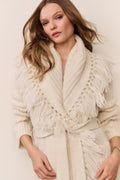 Woman wearing a cream-colored knitted cardigan with fringe details on a plain background