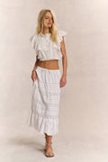 Woman wears white ruffle cropped top with midi white lace detail skirt against white background