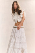 Woman wears white ruffle cropped top with midi white lace detail skirt and studded belt against white background