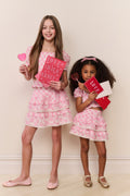 Two young girls in matching pink outfits holding heart-shaped cards against a light background