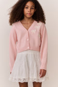 Tween wearing a pink sweater and white lace skirt on a plain background