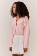 Tween wearing a pink sweater and white lace skirt on a plain background