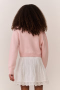 Tween wearing a pink sweater and white lace skirt on a plain background