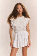 Woman wears white ruffle cropped top with mini white ruffle skirt against beige background