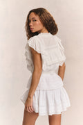 Woman wears white ruffle cropped top with mini white ruffle skirt against beige background