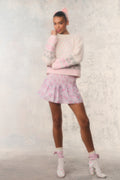 Woman wearing a light pink sweater and skirt with matching accessories on a textured white background