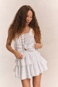 Woman wears white ruffle lace top with mini white ruffle skirt against beige background