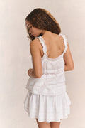 Woman wears white ruffle lace top with mini white ruffle skirt against beige background
