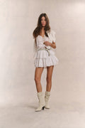 Woman wears white ruffle lace top with mini white ruffle skirt against beige background
