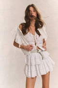 Woman wears white ruffle lace top with mini white ruffle skirt against beige background