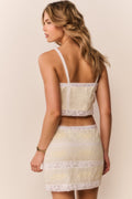 Woman wearing a white and yellow lace cropped top with matching skirt against a beige background
