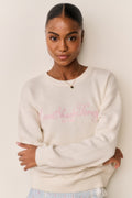 Woman wearing a cream sweater with pink LoveShackFancy text and a blue floral skirt against a beige background