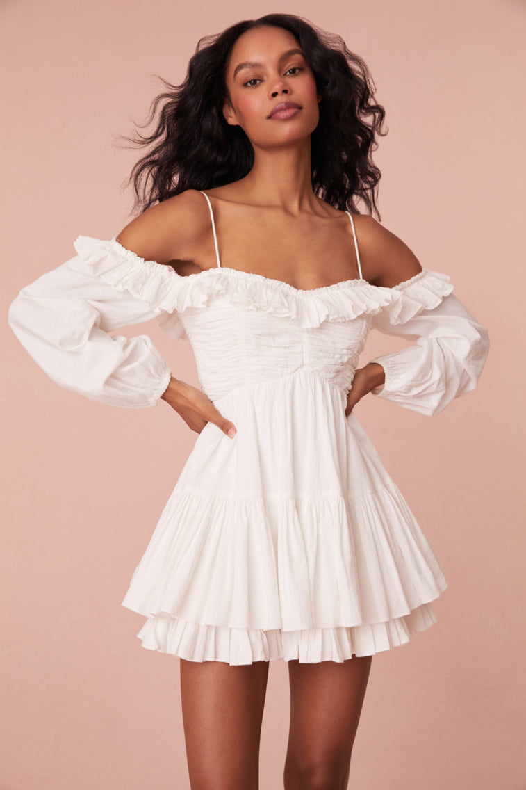White mini dress featuring a shaped waist with shirring and ruching at the bust. Includes off-shoulder blouson sleeves, elasticated openings, a two-tiered skirt, a smocked back waist, and adjustable spaghetti straps.
