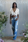 Woman wearing a white ruffled top and blue jeans with floral lace patterns, standing in front of a wooden wall with flowers.