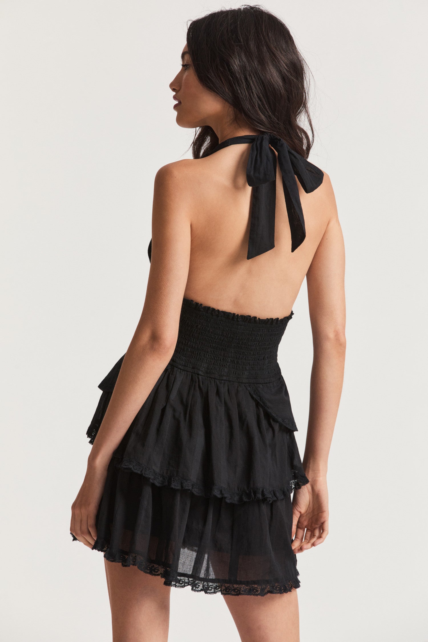Black mini dress with custom dobby striped cotton with all-over botanical embroidery. This low-cut convertible halter, with piping at the neckline, ties in a generous bow at the neck and can be crossed at front. Below a wide smocked waistband, the skirt flares to smaller details at the hips and two scalloped tiers with schiffli cutwork.