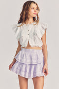 Woman wears white ruffle cropped top with mini purple tie-dye ruffle skirt against beige background