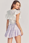 Woman wears white ruffle cropped top with mini purple tie-dye ruffle skirt against beige background