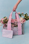 Floral-patterned transparent bag with pink straps held by a hand against a light blue background