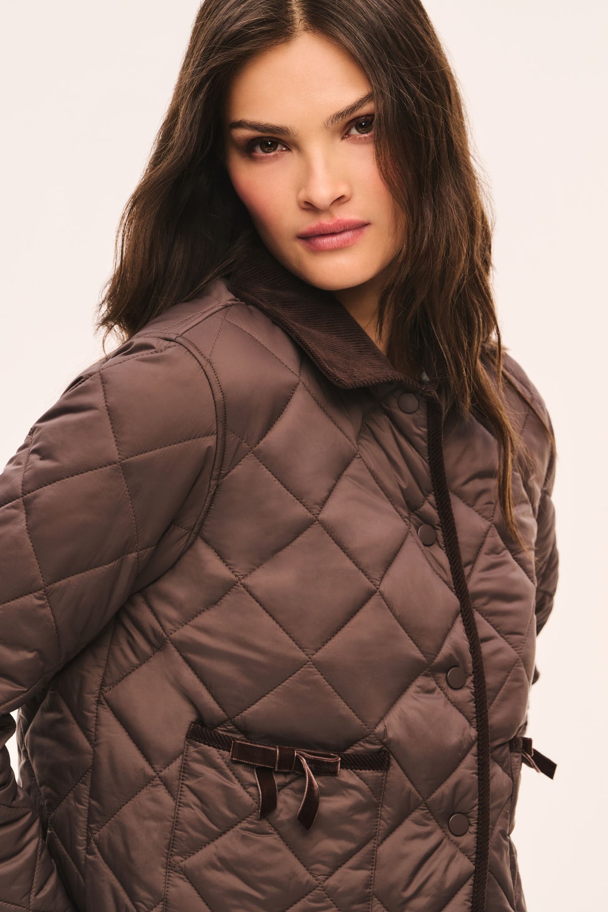 Close-up view of brown quilted jacket with dark brown corduroy collar and corduroy trim, snap button front, and front pockets with velvet bow trim