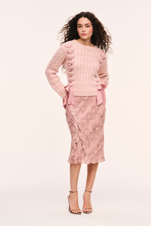 Pink textured knit pullover with crew neckline and ribbons laced down the front and bows at the hem, paired with pink snakeskin print skirt