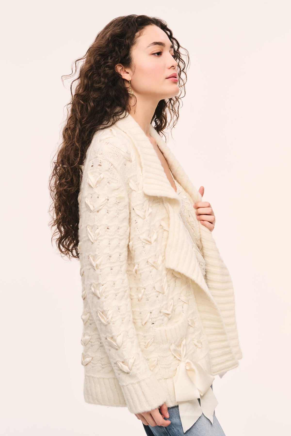 Windel ivory knit cardigan side view: ivory textured knit cardigan, open front with shawl collar, ribbon laced through the front, and ribbon bows on the pockets