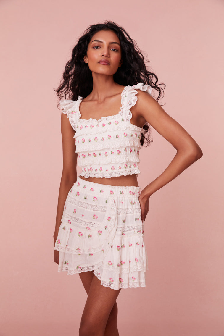 Top embroidered with small pink roses, the piece begins with tiny flutter sleeves with elastic at the shoulders above a square neckline. Smocked all over, the top includes lace and ruffle details throughout and stops at the natural waist.