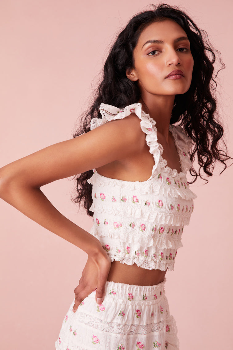 Top embroidered with small pink roses, the piece begins with tiny flutter sleeves with elastic at the shoulders above a square neckline. Smocked all over, the top includes lace and ruffle details throughout and stops at the natural waist.