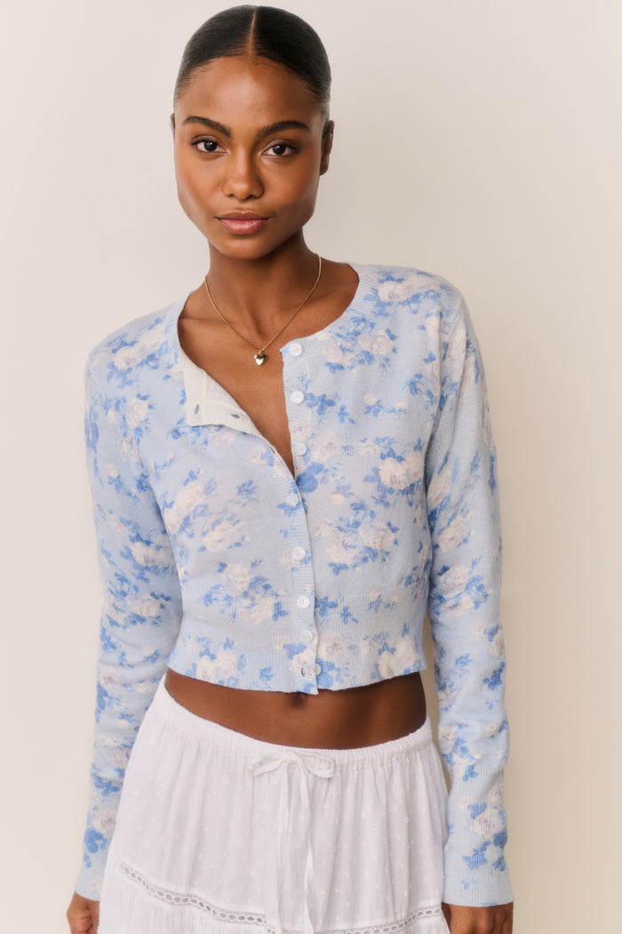 Woman wearing a light blue floral cardigan over a white top and skirt on a beige background