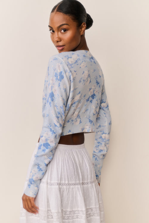 Woman wearing a light blue floral cardigan over a white top and skirt against a beige background