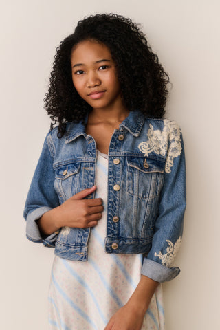 Young girl wearing a denim jacket with lace embroidery against a plain background