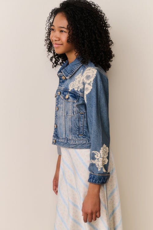 Young girl wearing a denim jacket with lace embroidery against a plain background