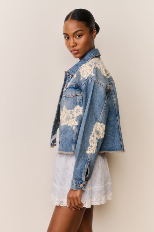 Woman wears blue denim jacket with floral lace embroidery and a light blue floral skirt on a beige background