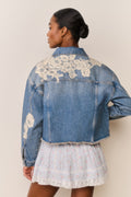 Woman wears blue denim jacket with floral lace embroidery and a light blue floral skirt on a beige background
