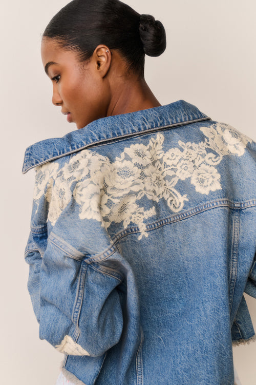 Woman wears blue denim jacket with floral lace embroidery and a light blue floral skirt on a beige background
