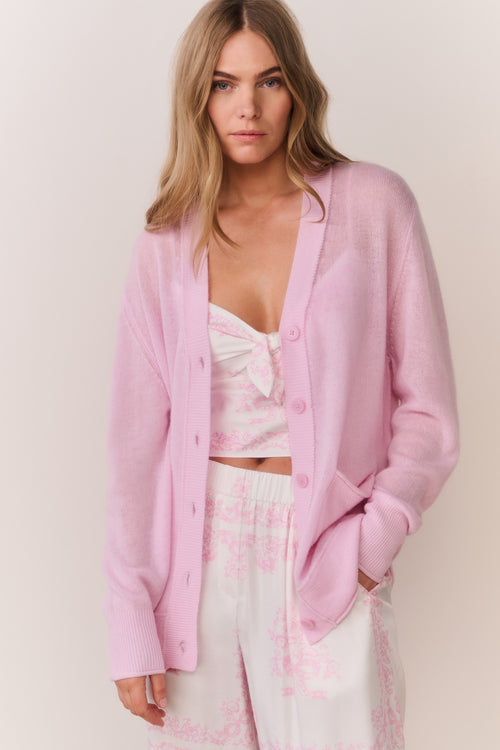 Pink cardigan worn by a woman in pink and white patterned pants and top on a plain background