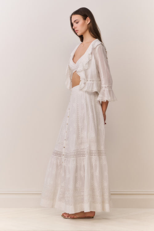 Woman wearing a white button down blouse with ruffled details and bralette top with matching maxi skirt on a plain background 