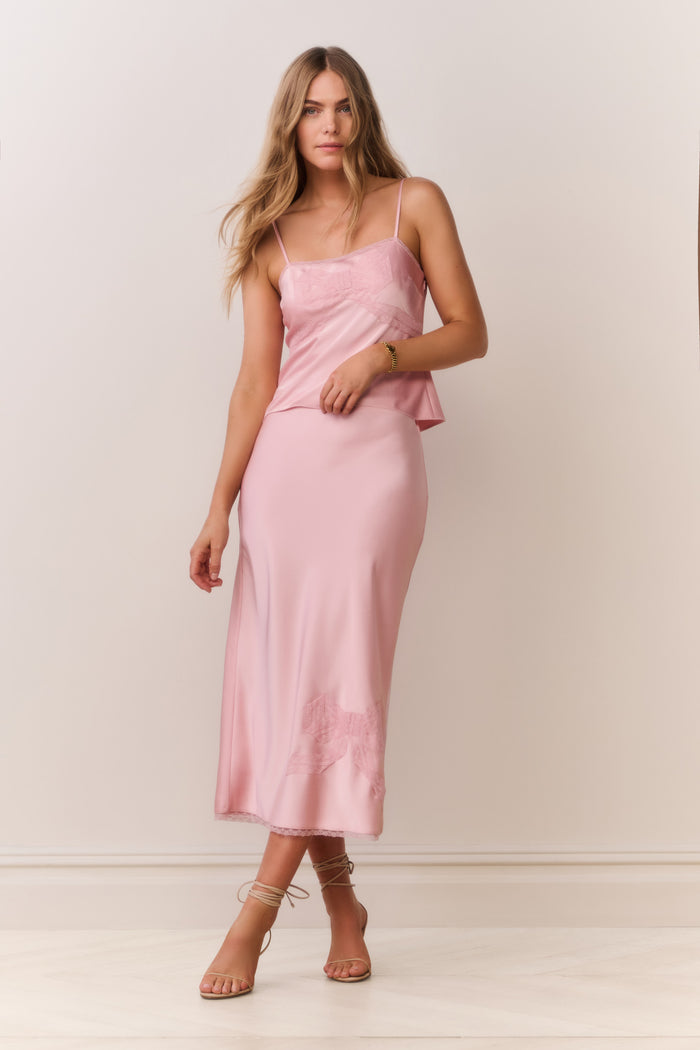 Woman wearing a pink satin cami and matching skirt with thin straps and bow detail on a plain background
