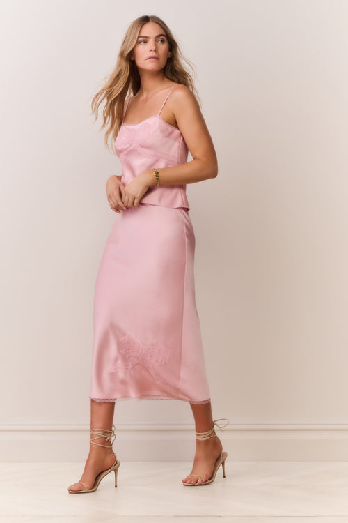 Woman wearing a pink satin cami and matching skirt with thin straps and bow detail on a plain background
