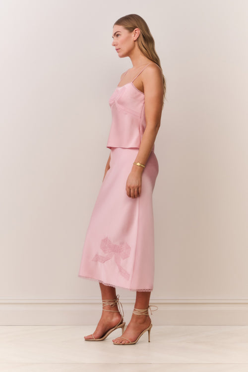 Woman wearing a pink satin cami and matching skirt with thin straps and bow detail on a plain background