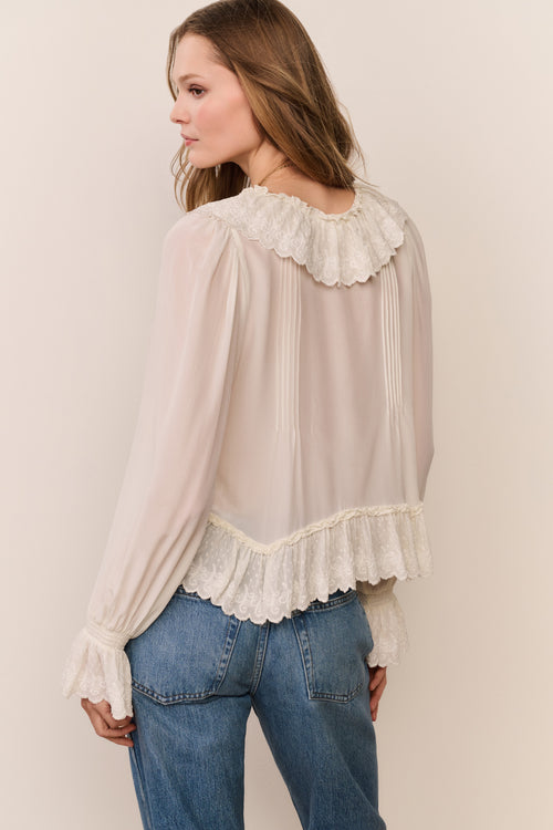 Woman wearing a beige blouse with lace details and blue jeans against a plain background