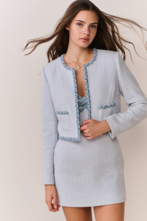 Woman wearing a light blue tweed jacket with decorative trim and matching mini dress on a plain background